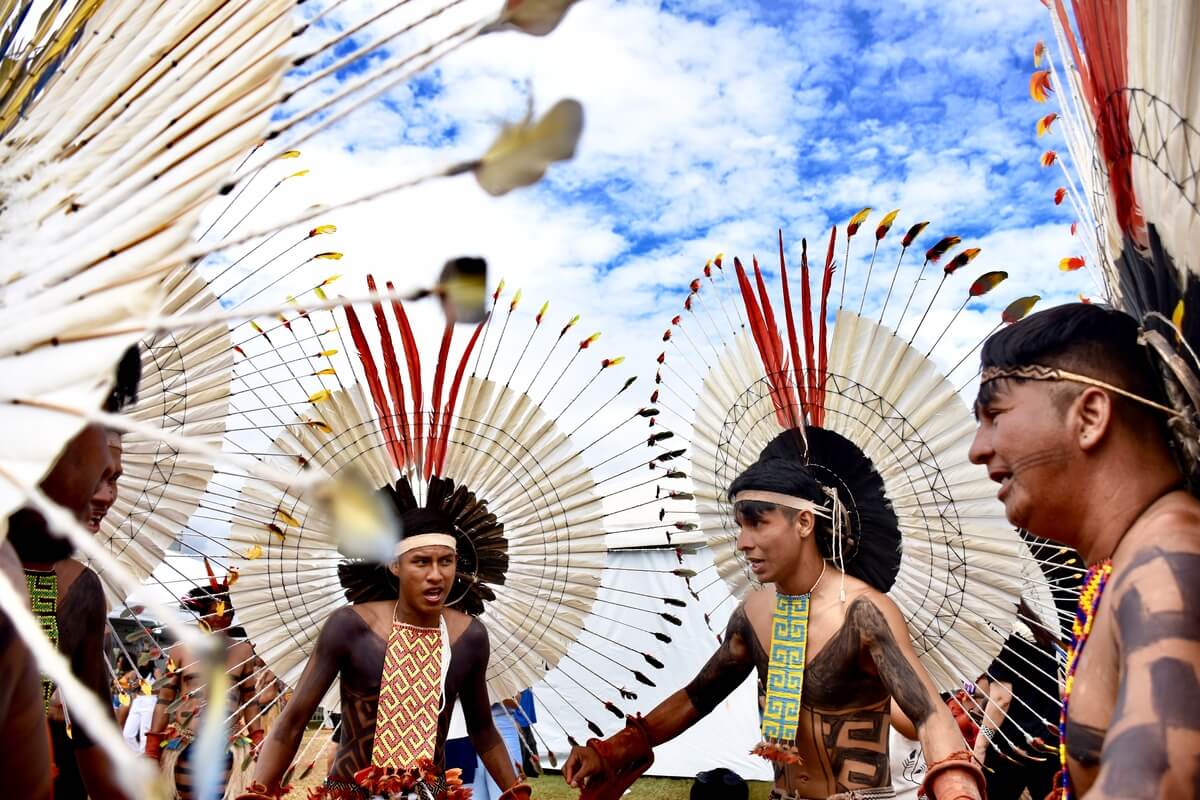 Imagem mostra quatro homens indígenas caracterizados com adereços e pinturas tradicionais em roda