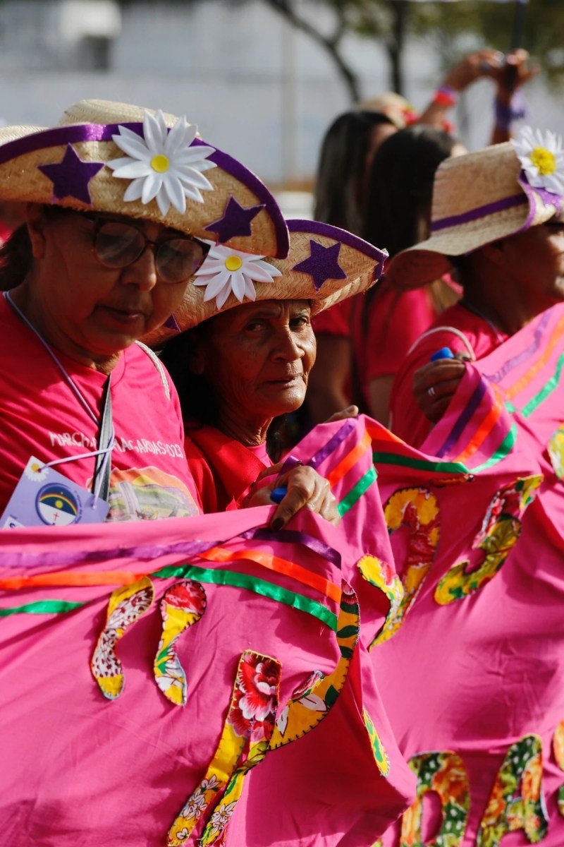 Mulheres vestindo rosa e usando chapéus de cangaceiro com flores seguram tecido rosa com retalalhos coloridos